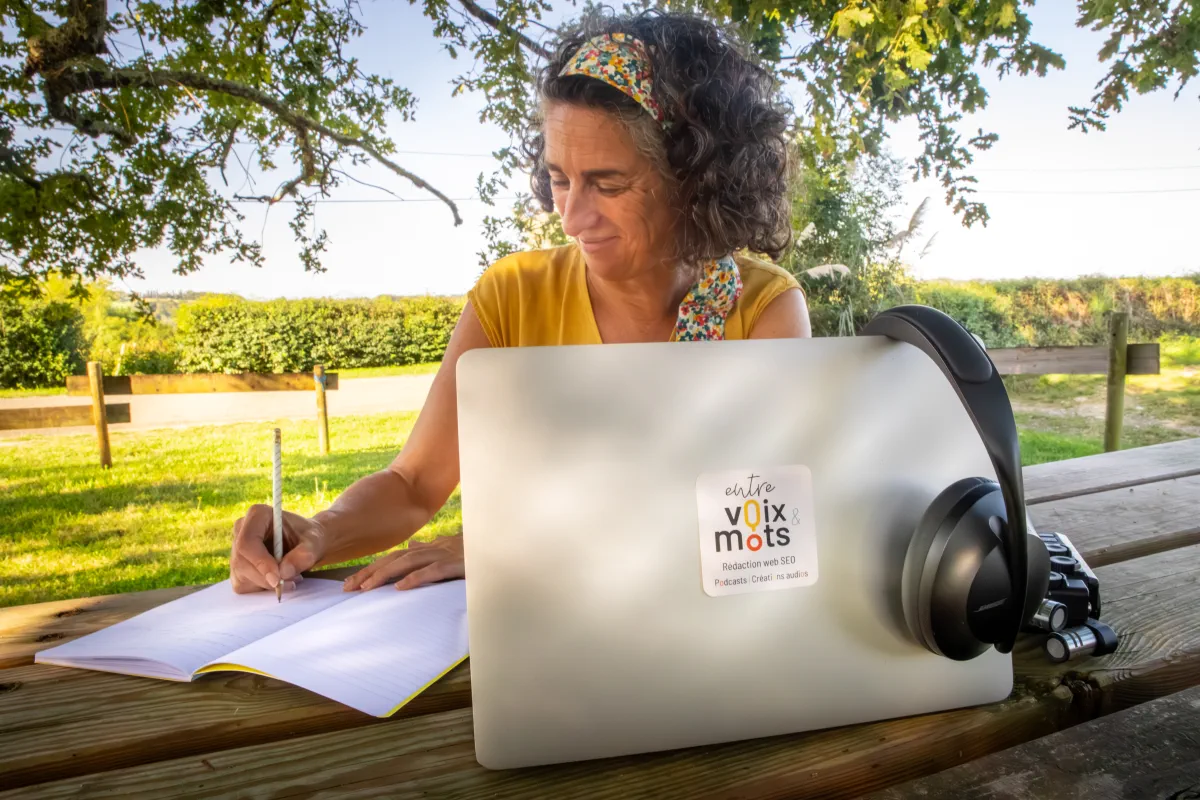 Portrait de Christine Irabola qui écrit dans un carnet avec ordinateur et matériel de podcast à proximité